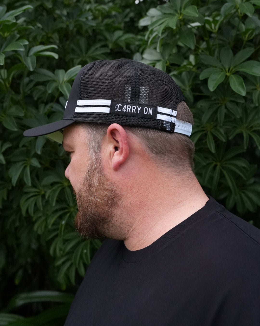 Person wearing a black cap with text, standing among green foliage