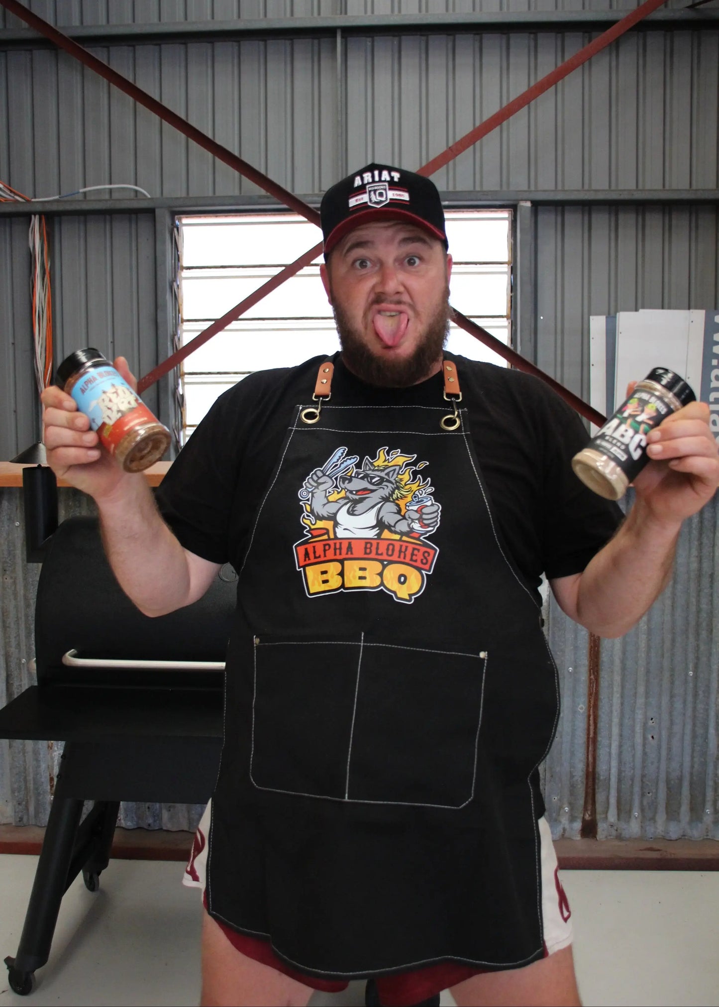 Man wearing a black apron with a BBQ logo, holding two rubs, in an industrial setting.